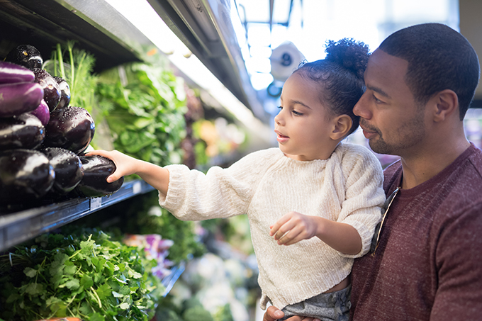 A pre-school age girl helping her dad pick out veggies in the produce section at the grocery store.