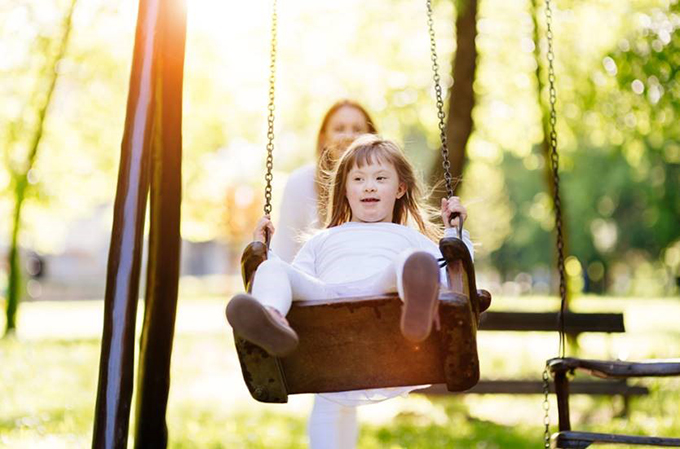 A girl with Down Syndrome smiling on a swing in a park.